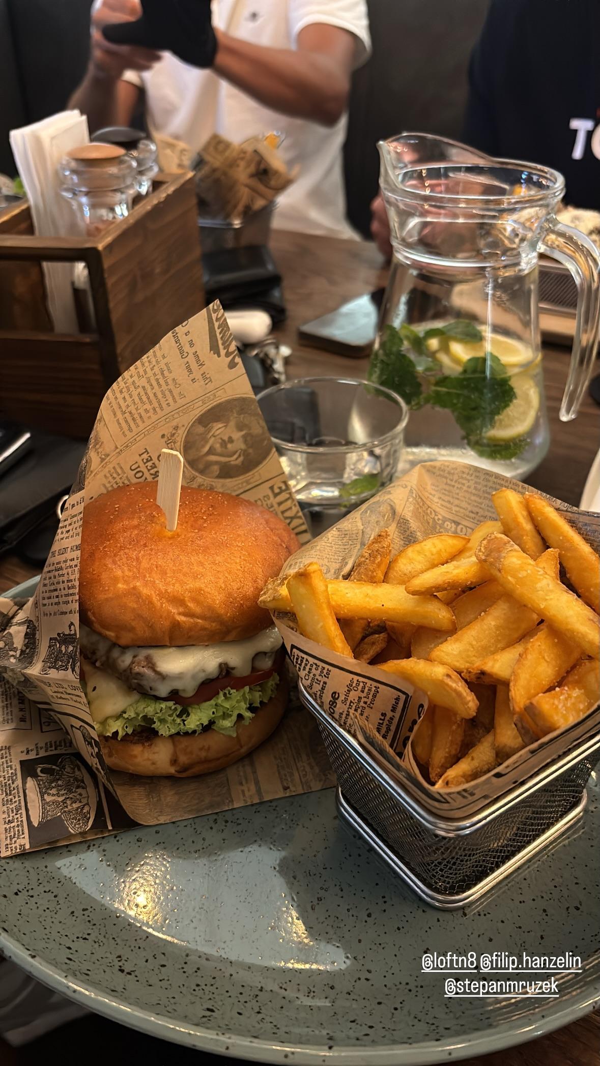 Instagram story from Loft N8: a cheeseburger and a basket of fries served on newspaper paper, next to a jug of lemon and mint water