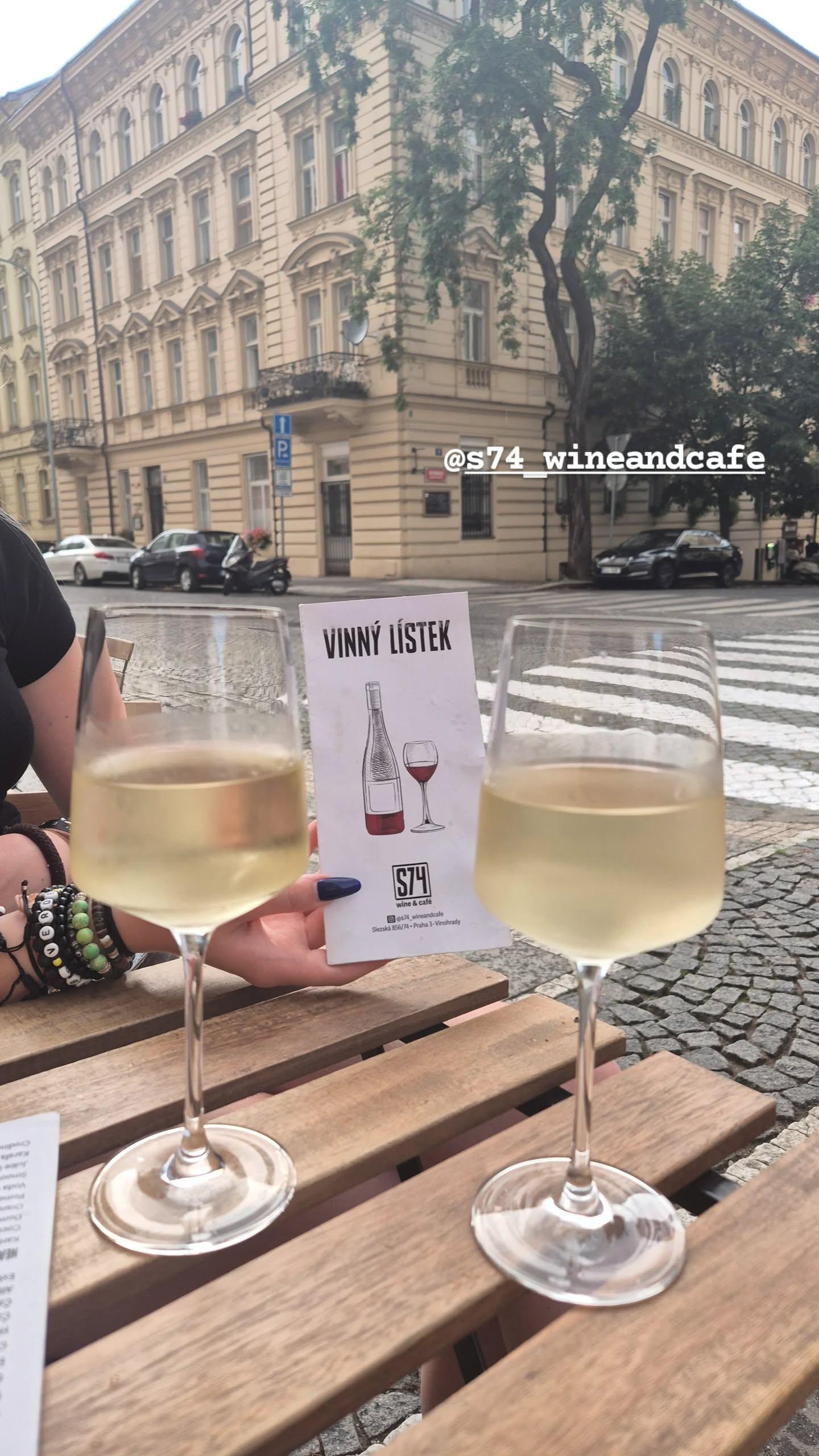 Instagram story from S74 wine&café: two glasses of white wine on an outdoor table with the wine list held up, on a Prague street corner